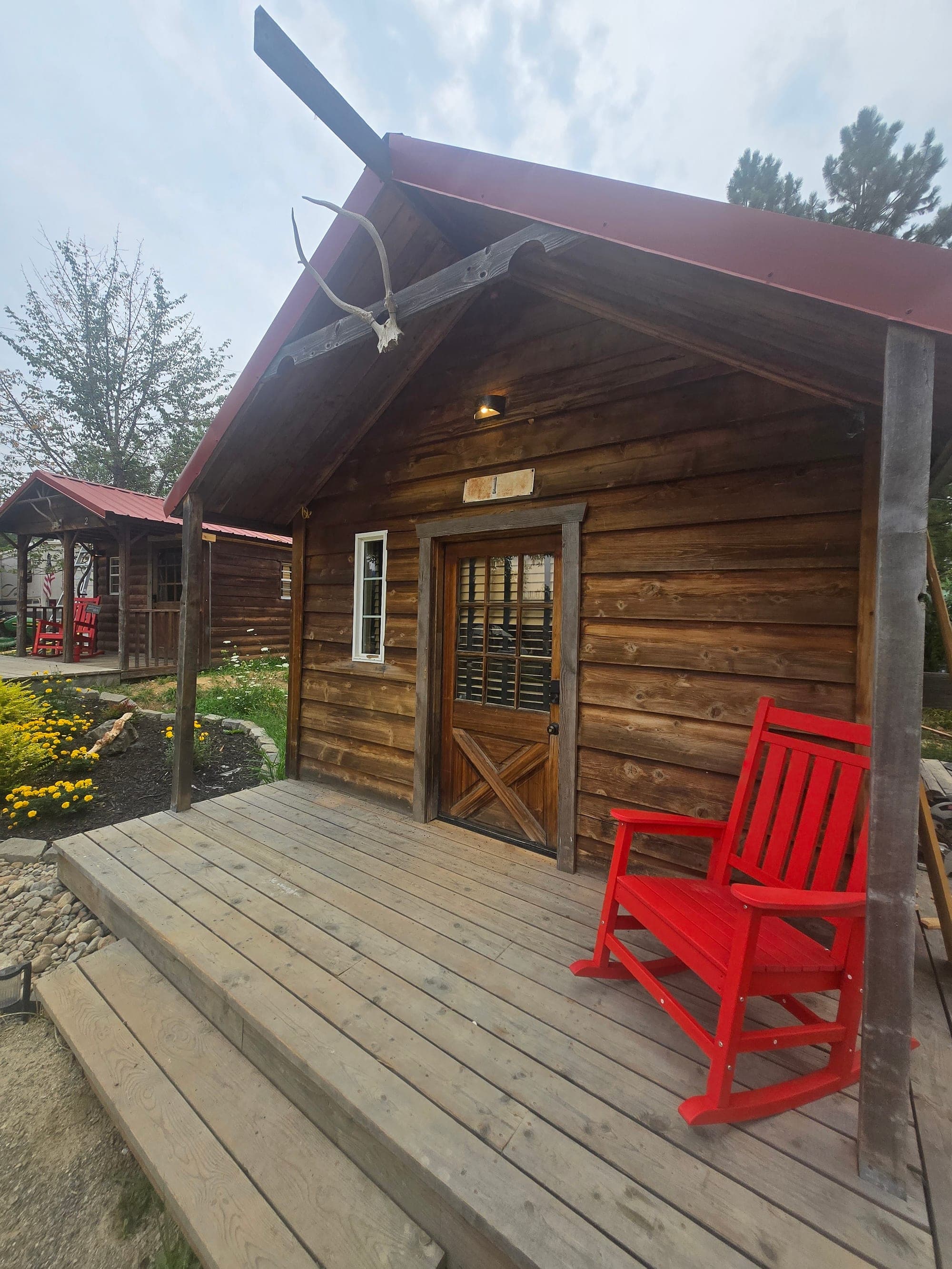 Log cabin front porch with red rocker, antlers above the door, and a second cabin in the distance