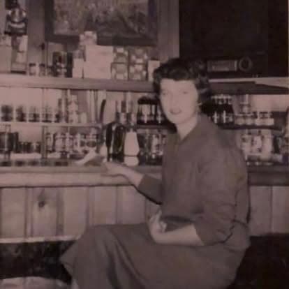 Sepia-toned vintage photograph of a woman seated inside the historic tavern with stocked general-store shelves behind her